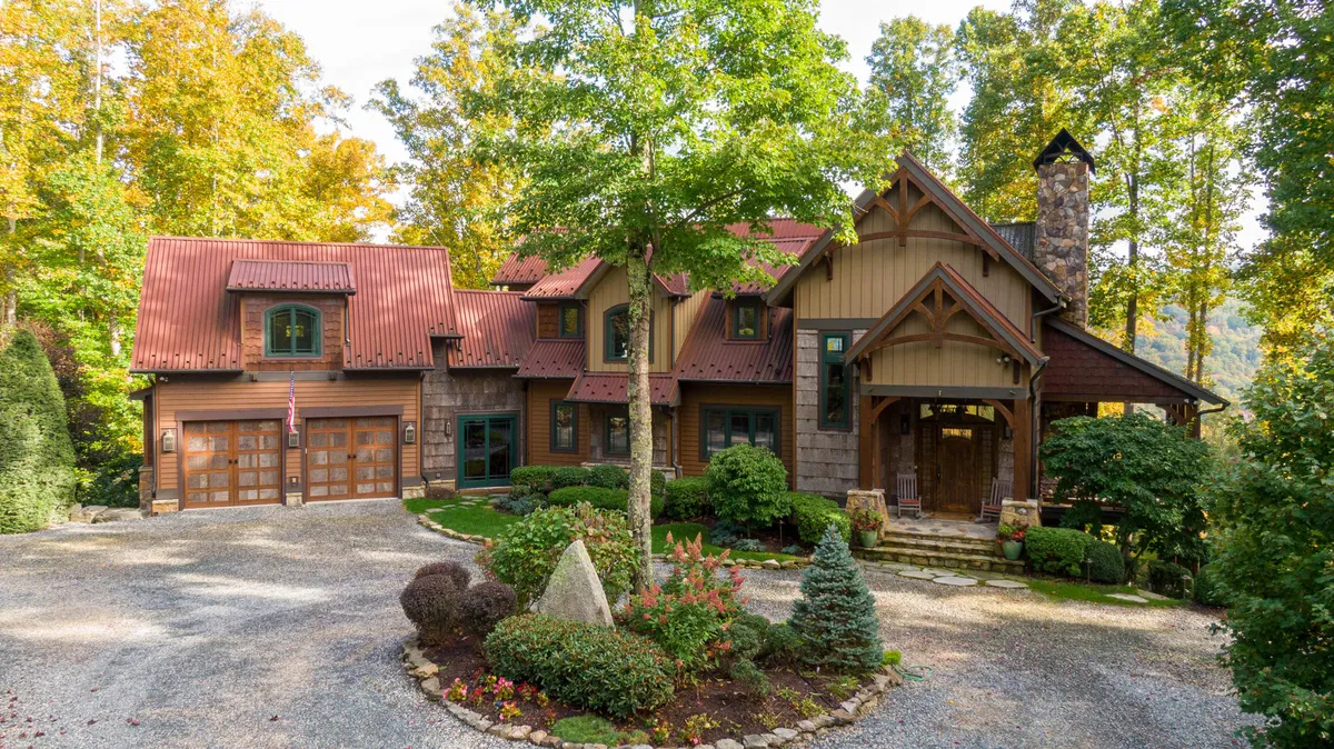 The hero image showcases the grand front entrance of a custom-built timber-framed mountain lodge, featuring a majestic stone chimney, beautifully landscaped gardens, and expansive decks with rustic branch railings. The setting is serene, wrapped in natural beauty, inviting guests to imagine warm sunset gatherings or tranquil mornings with coffee on the porch.
