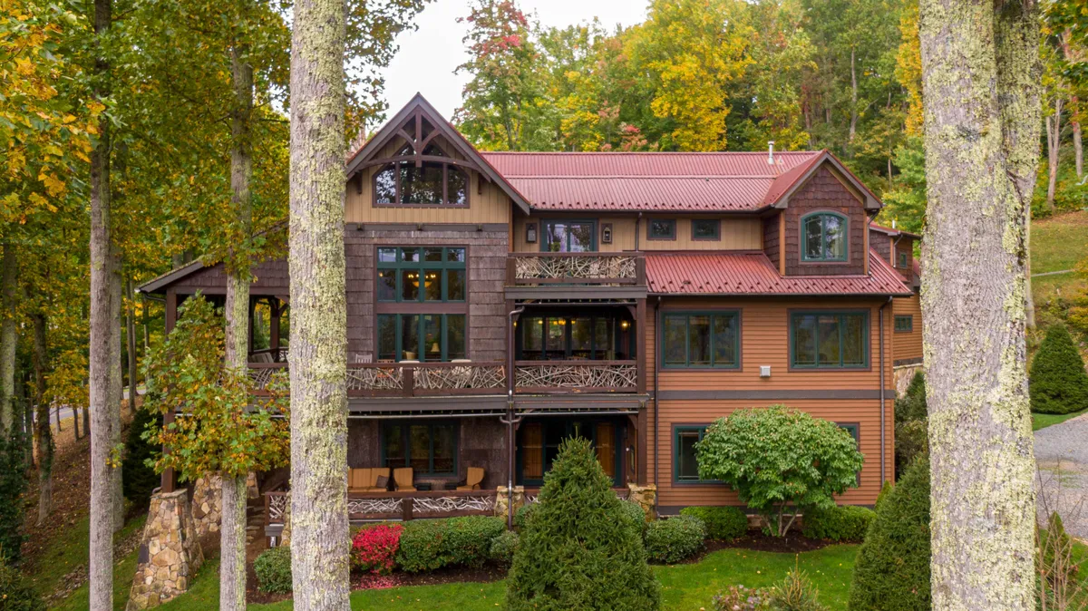 A striking exterior view of the large wooden structure surrounded by mature trees and colorful landscaping, with ornate balcony railings. The porch and balcony areas are visible, inviting outdoor gatherings or relaxed moments overlooking the scenery.