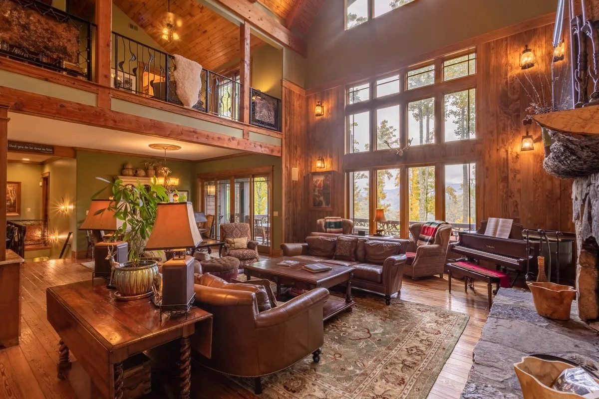 A stunning Great Room with expansive windows framing breathtaking mountain vistas. The plush, arranged seating area centers around a grand stone fireplace, while a Baby Grand Piano stands ready for gatherings. The room is illuminated by warm natural light, with inviting textures and a harmonious blend of timber and stone elements, reflecting the lodge's luxurious yet comfortable spirit.