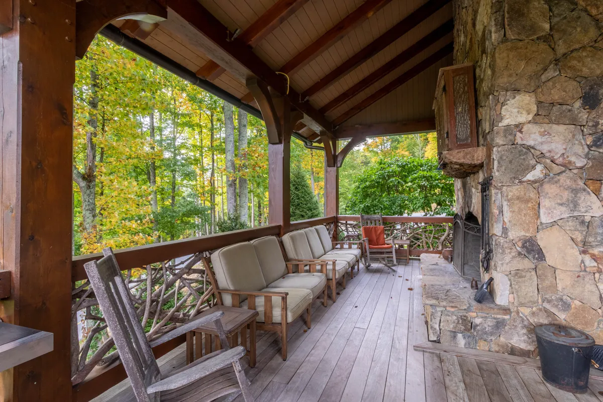 This exterior shot features wooden chairs positioned around a commanding stone fireplace, set against a backdrop of lush green foliage. The scene evokes the ambiance of an outdoor living area designed for relaxation and gathering, with the stone fireplace as the centerpiece.