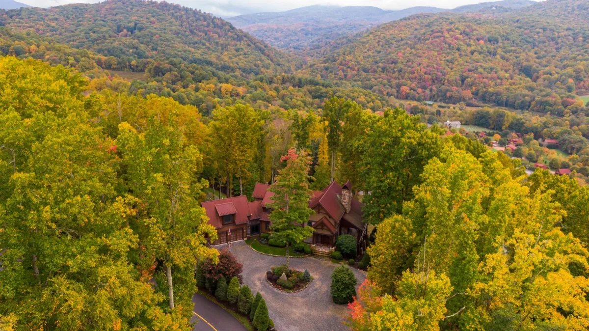This exterior image features the main house set against a backdrop of mature trees, with a winding driveway leading up to the property. The surrounding natural landscape hints at woodland trails and the spaciousness of the grounds, setting an inviting, tranquil mountain scene.