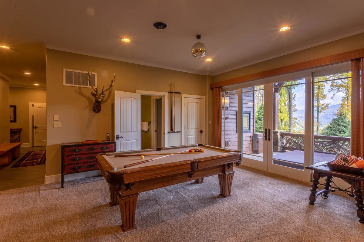 A bright, playful room with a prominently featured pool table set against large windows, rustic decor, and views out to the surrounding landscape. The space feels open and social, ideal for friendly competitions or relaxed gatherings.